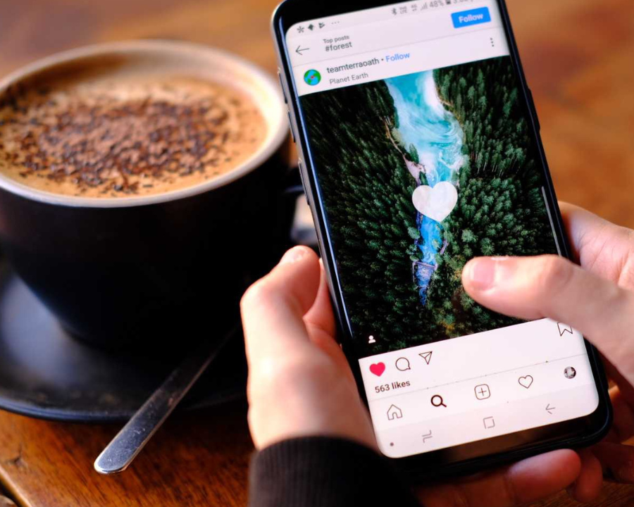 Eine Person scrolls am Handy durch Instagram und likes Bilder. Eine Tasse Kaffee im Hintergrund.