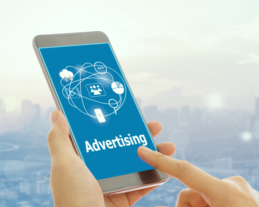 Person hält ein Smartphone in der Hand, auf dem der Schriftzug „Advertising“ sowie verschiedene Symbole für digitale Werbung (wie Diagramme, Netzwerkverbindungen und Nutzer-Icons) angezeigt werden; im Hintergrund eine verschwommene Stadtansicht.