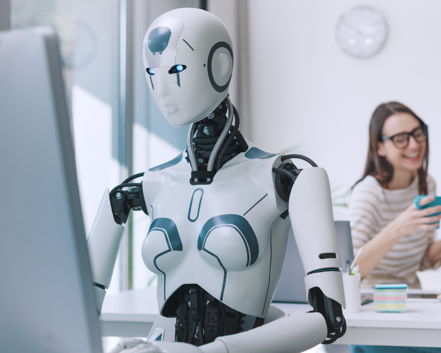 Ein humanoider Roboter mit weiblichen Merkmalen arbeitet konzentriert an einem Computer in einem modernen Büro, im Hintergrund sitzt eine lachende Frau mit Smartphone.